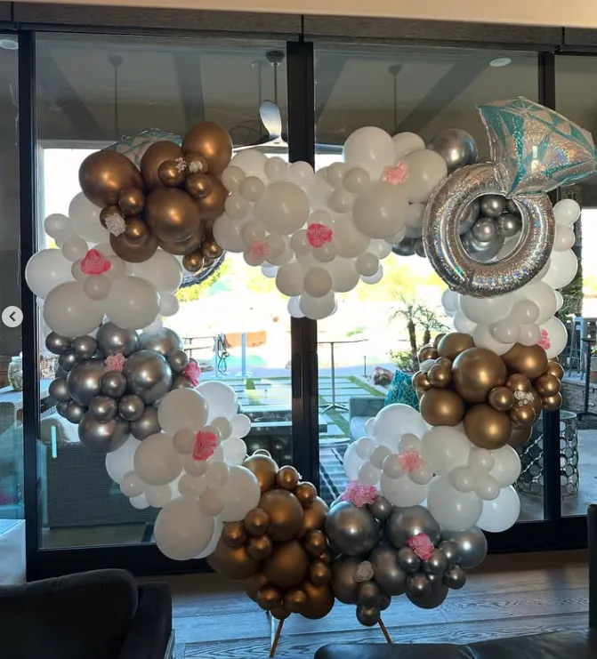 Heart-shaped balloon arch in white, gold, silver, and pink, displayed in front of glass doors.