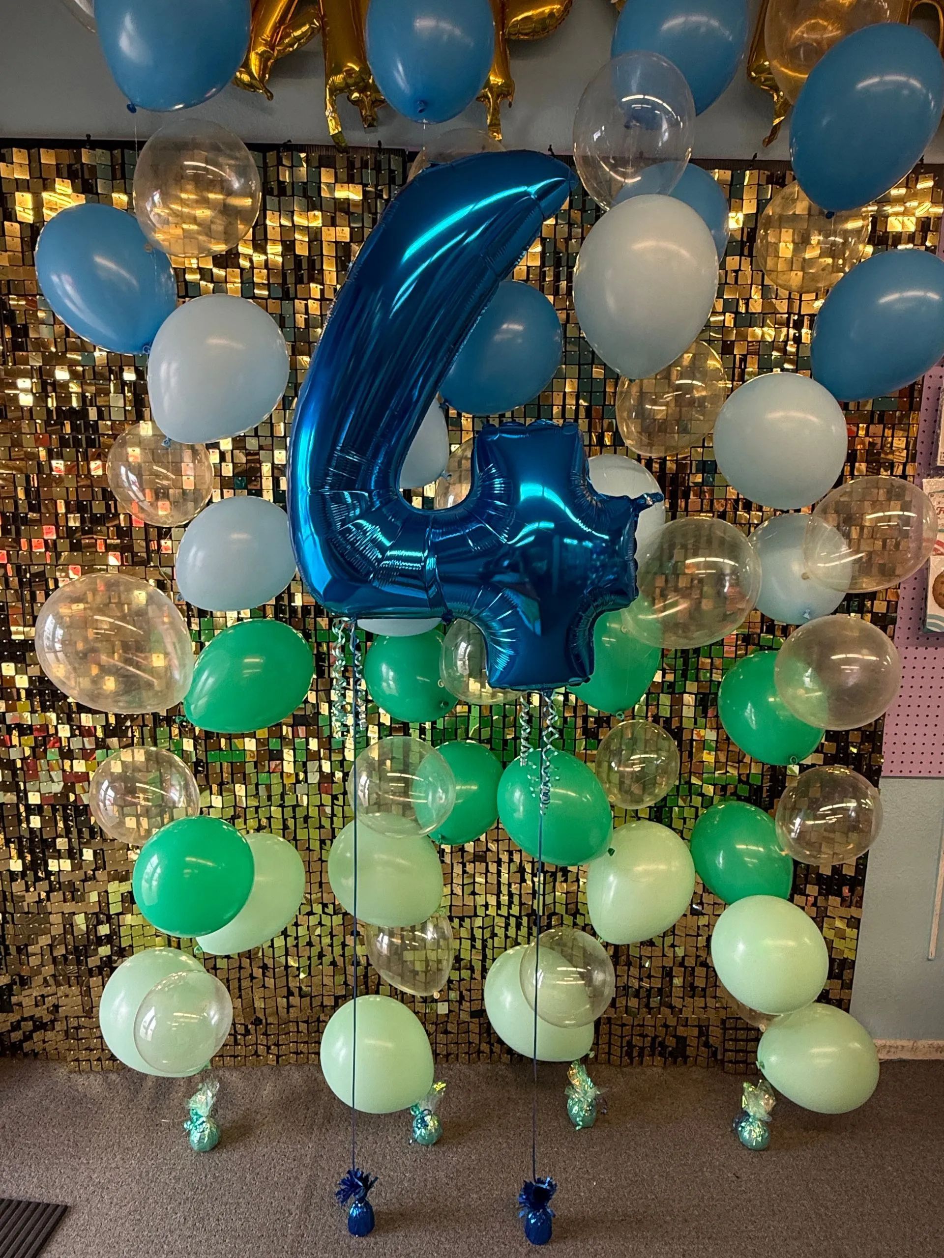 Blue metallic number 4 balloon amid blue, white, and green balloons on a gold sequin backdrop