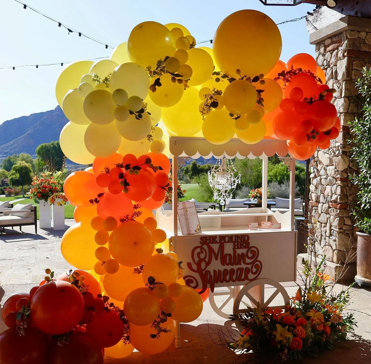 Colorful balloon arch in yellow, orange, and red framing a rustic cart at an outdoor event