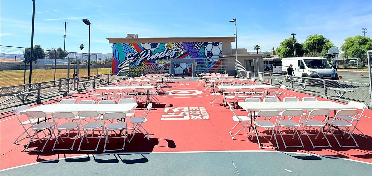 Outdoor dining setup with white tables and red flooring before a colorful mural wall under a clear sky