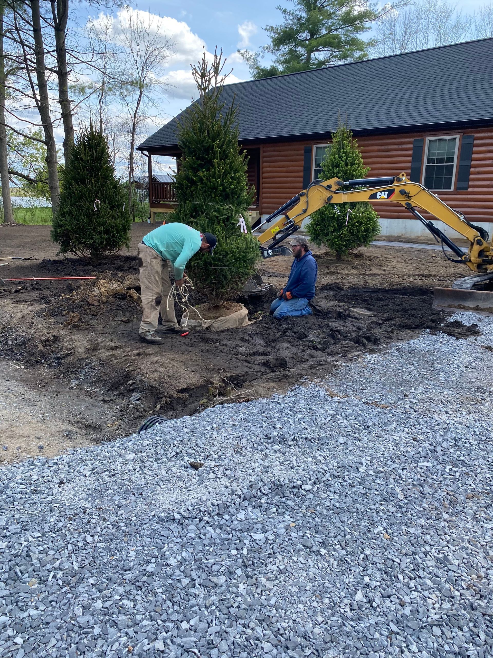 Two people working on a landscaping project next to a gravel driveway near a log cabin; one digs, one kneels.