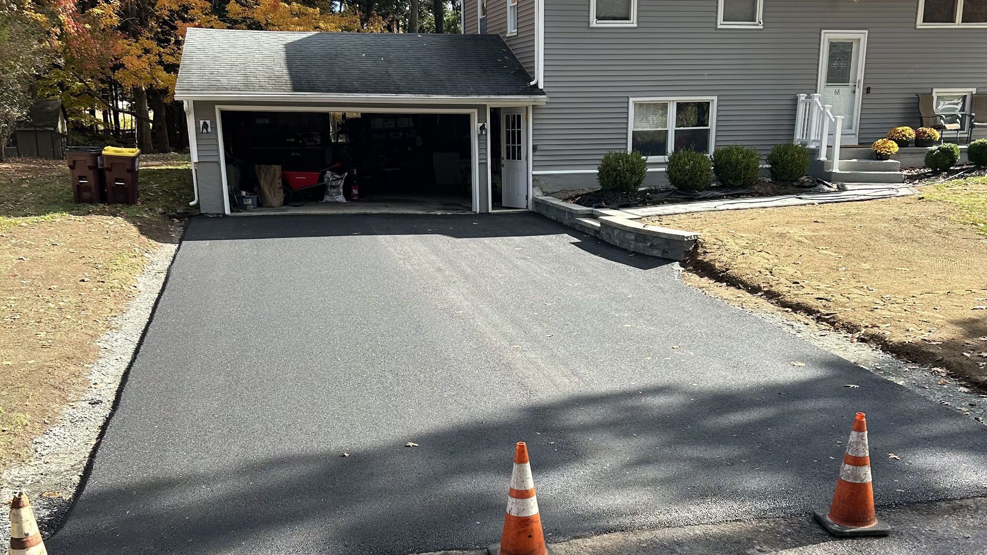 Newly paved asphalt driveway leading to a gray house with garage. Orange cones present. Fall foliage.