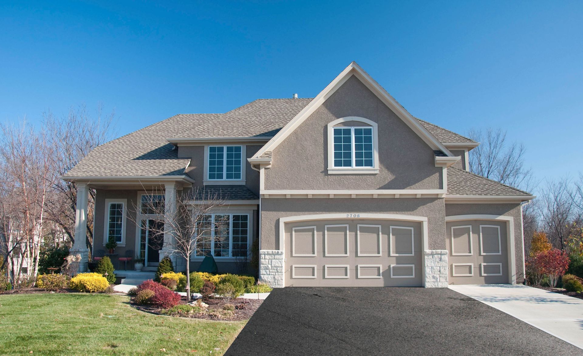 Two-story house with tan stucco exterior, gray roof, and two-car garage. Driveway leads to the house, clear blue sky.