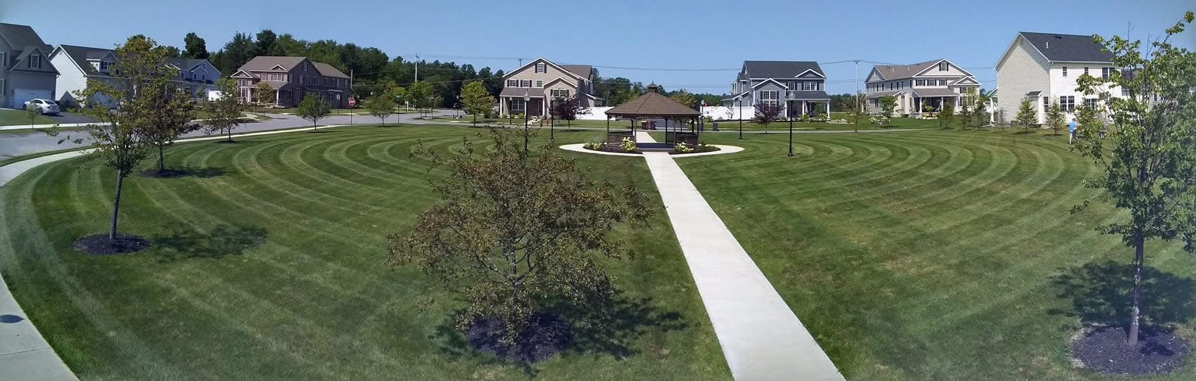A well-manicured lawn with a walking path, small gazebo, and houses on a sunny day.