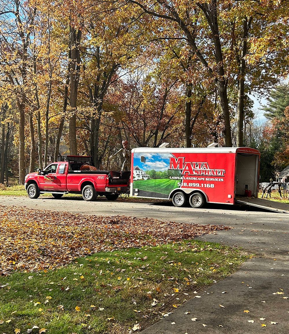 Red truck towing a red trailer, both with landscaping business logos, on a driveway with fall leaves.
