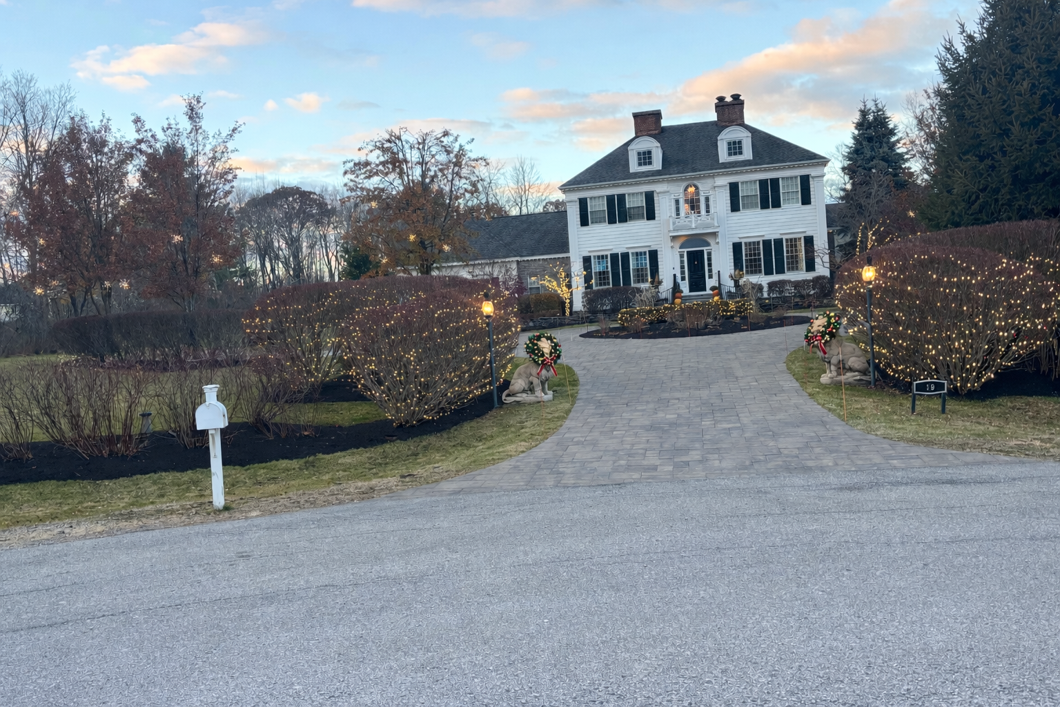 White colonial house with holiday lights; gravel driveway.
