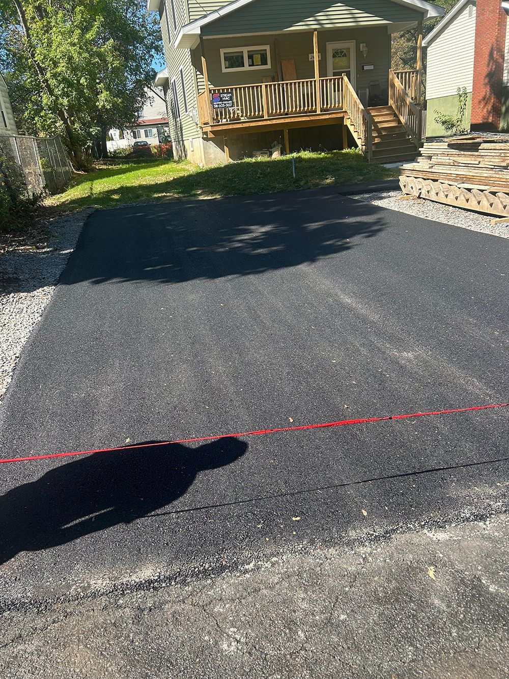 Newly paved asphalt driveway leading to a beige house with a dark brown garage door, trees in background.
