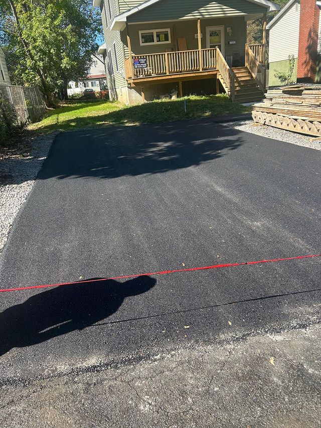 Newly paved asphalt driveway leading to a beige house with a dark brown garage door, trees in background.