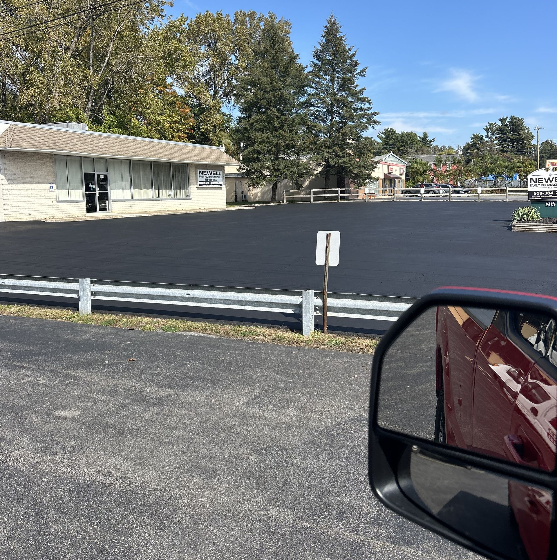 Newly paved parking lot with a building and a vehicle's side mirror visible. Blue sky, fall foliage.