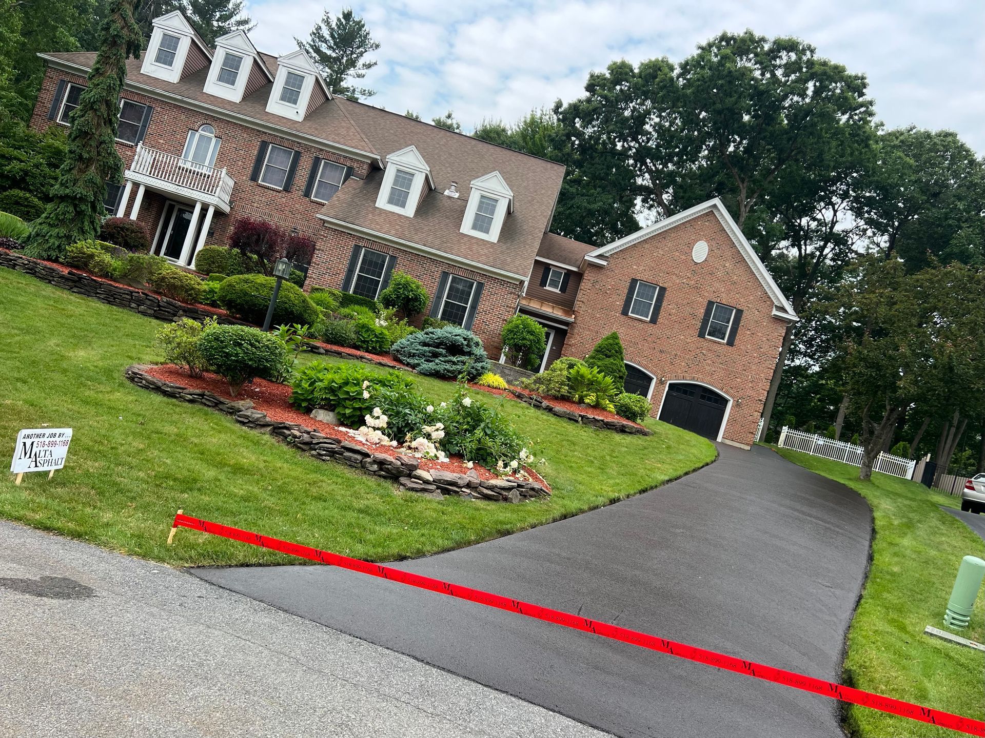 Two-story brick house with a sloped lawn and black driveway. Landscaping features red mulch. Overcast sky.
