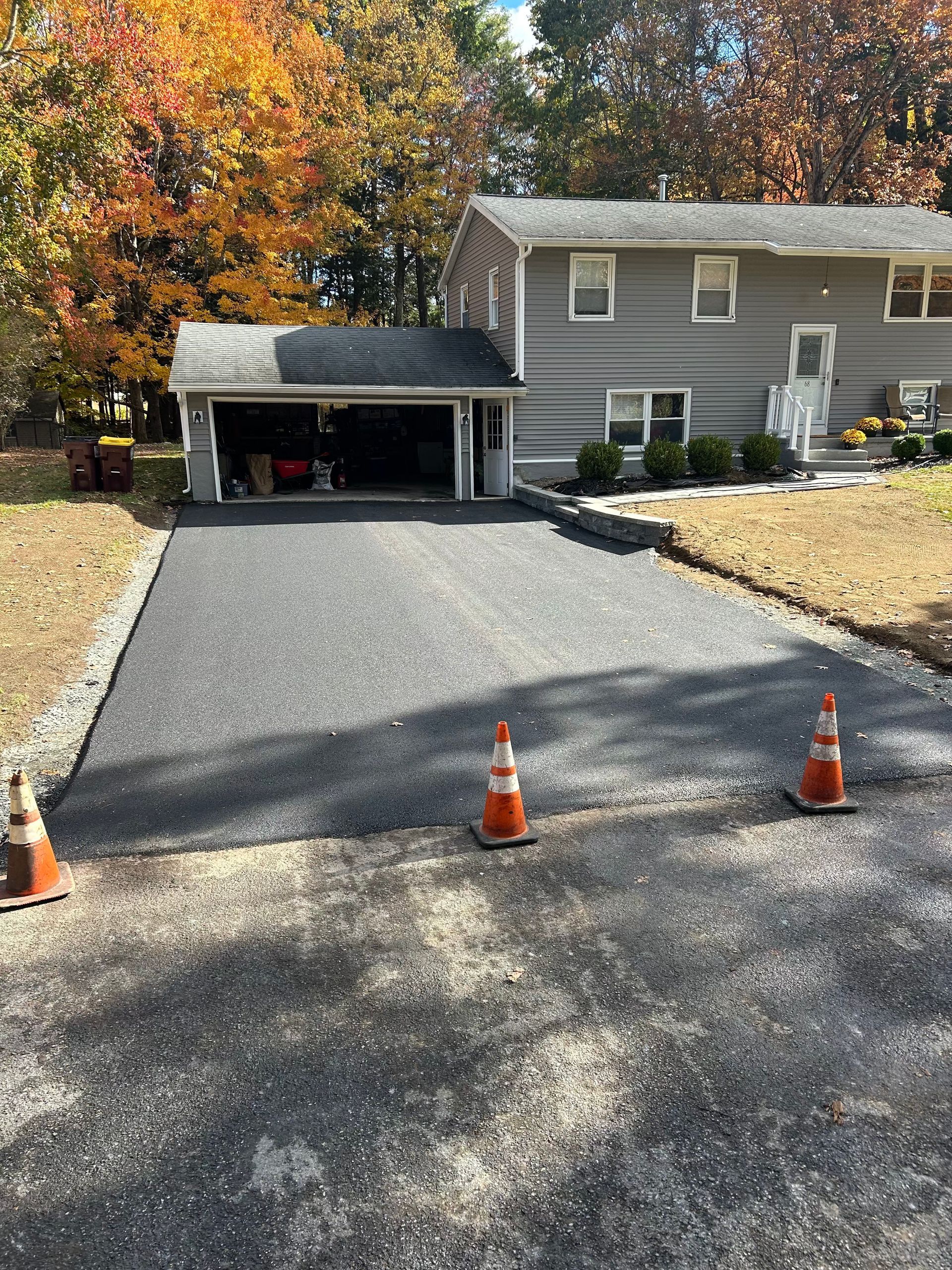Newly paved asphalt driveway leading to a two-story gray house with garage; orange traffic cones in foreground.
