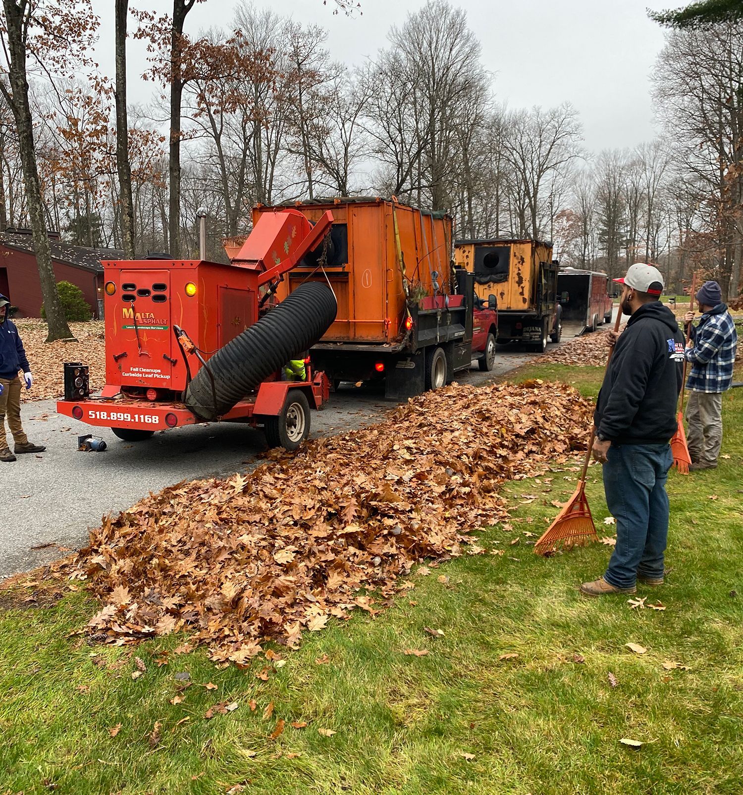 Leaf removal operation: orange leaf shredder, truck, workers, piles of leaves, grassy area, paved driveway.