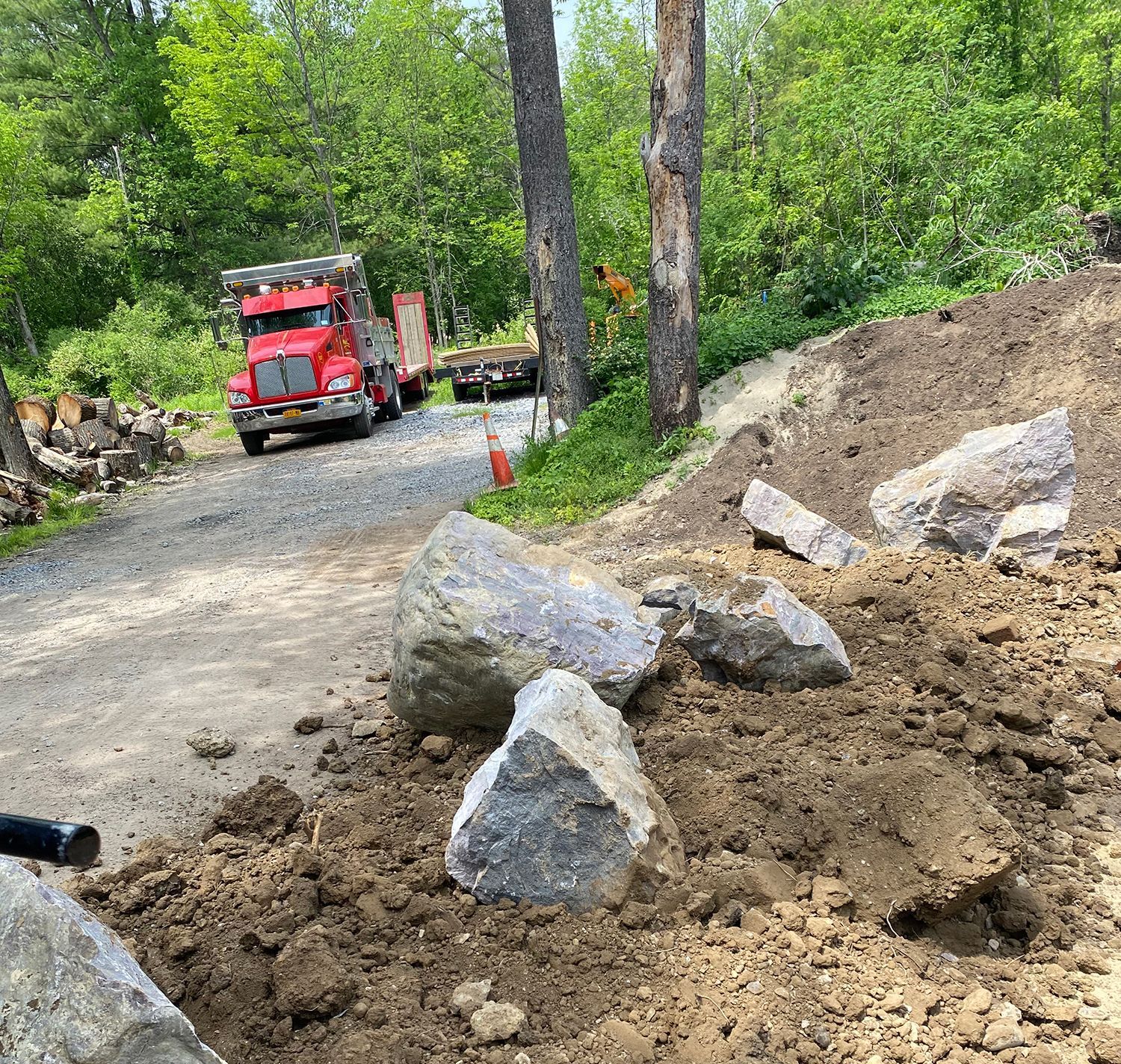 Red truck unloading large rocks on dirt road with trees in the background.