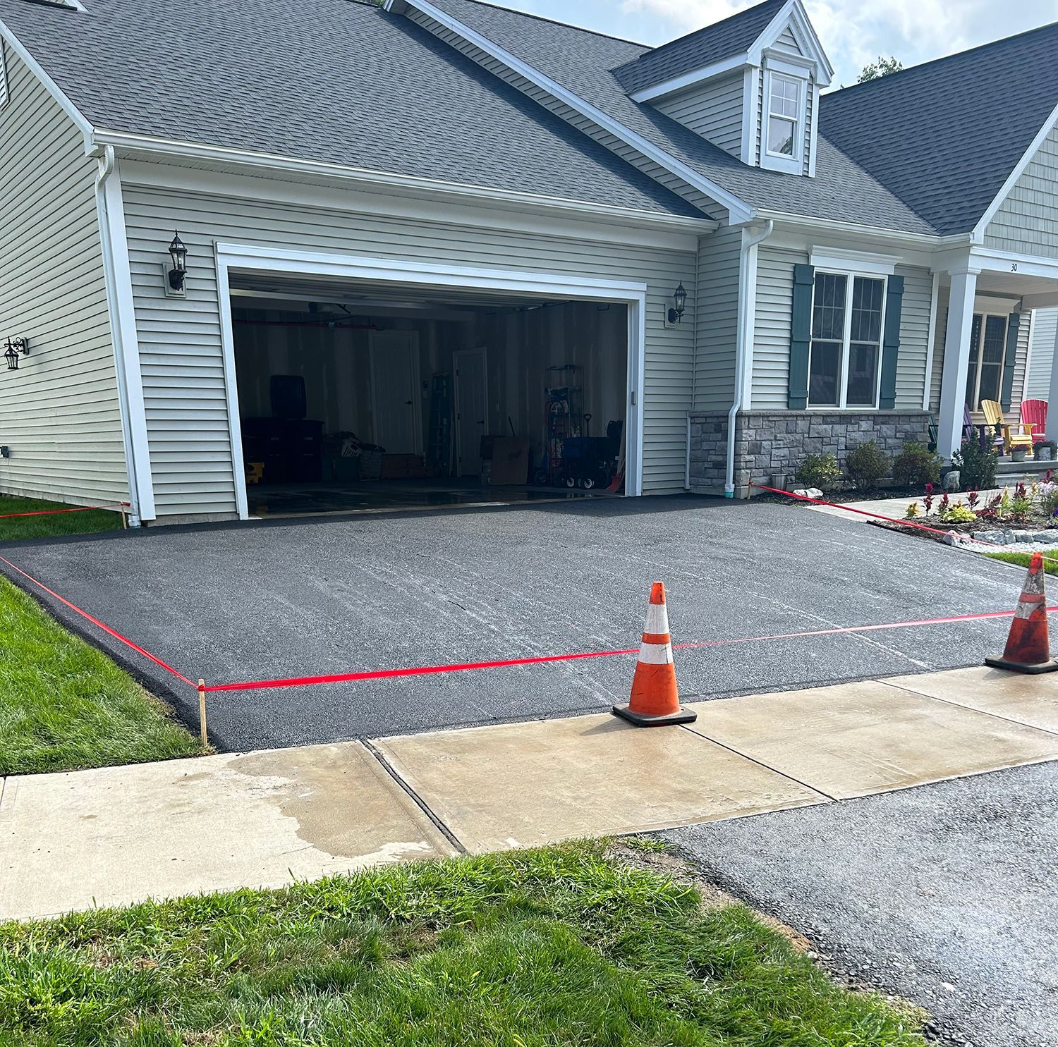 New asphalt driveway in front of a house, with orange cones, red tape, and an open garage.