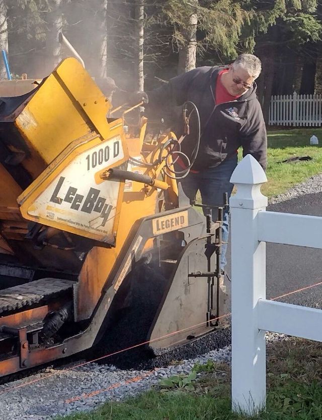 Man operating a yellow asphalt paving machine, laying asphalt on a road.