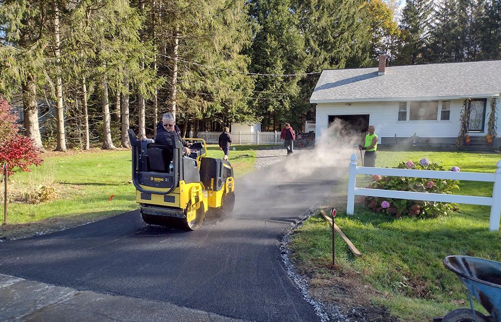Asphalt paving in progress on a driveway. A man operates a yellow roller; workers nearby. House and trees in the background.