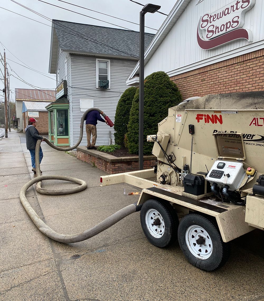 Two people using a hose connected to a machine on a trailer, near a building.