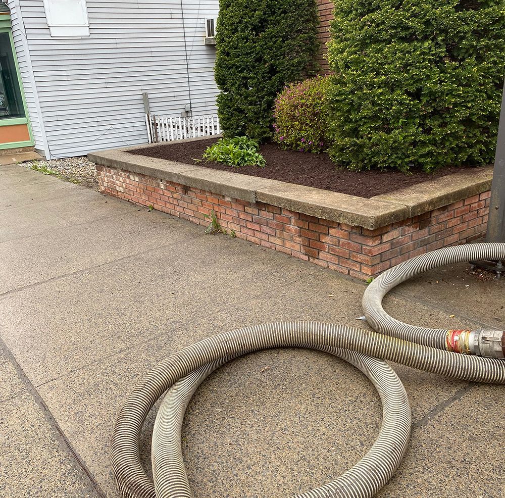 A brick planter box with shrubs sits next to a sidewalk and coiled hose. A building is behind it.