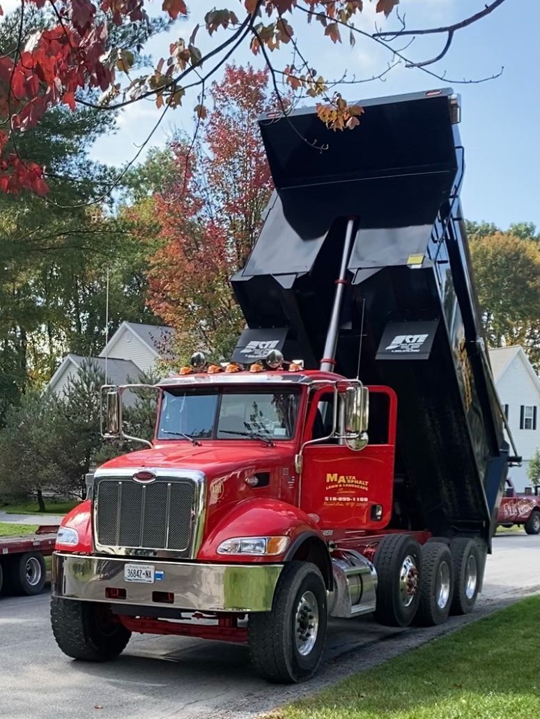 Red dump truck with raised black bed on a residential street. Fall foliage in the background.