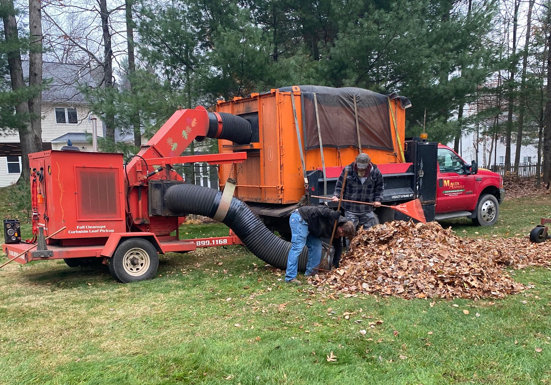 Two people loading leaves into a leaf vacuum truck on a lawn. Orange and red machine; fall setting.