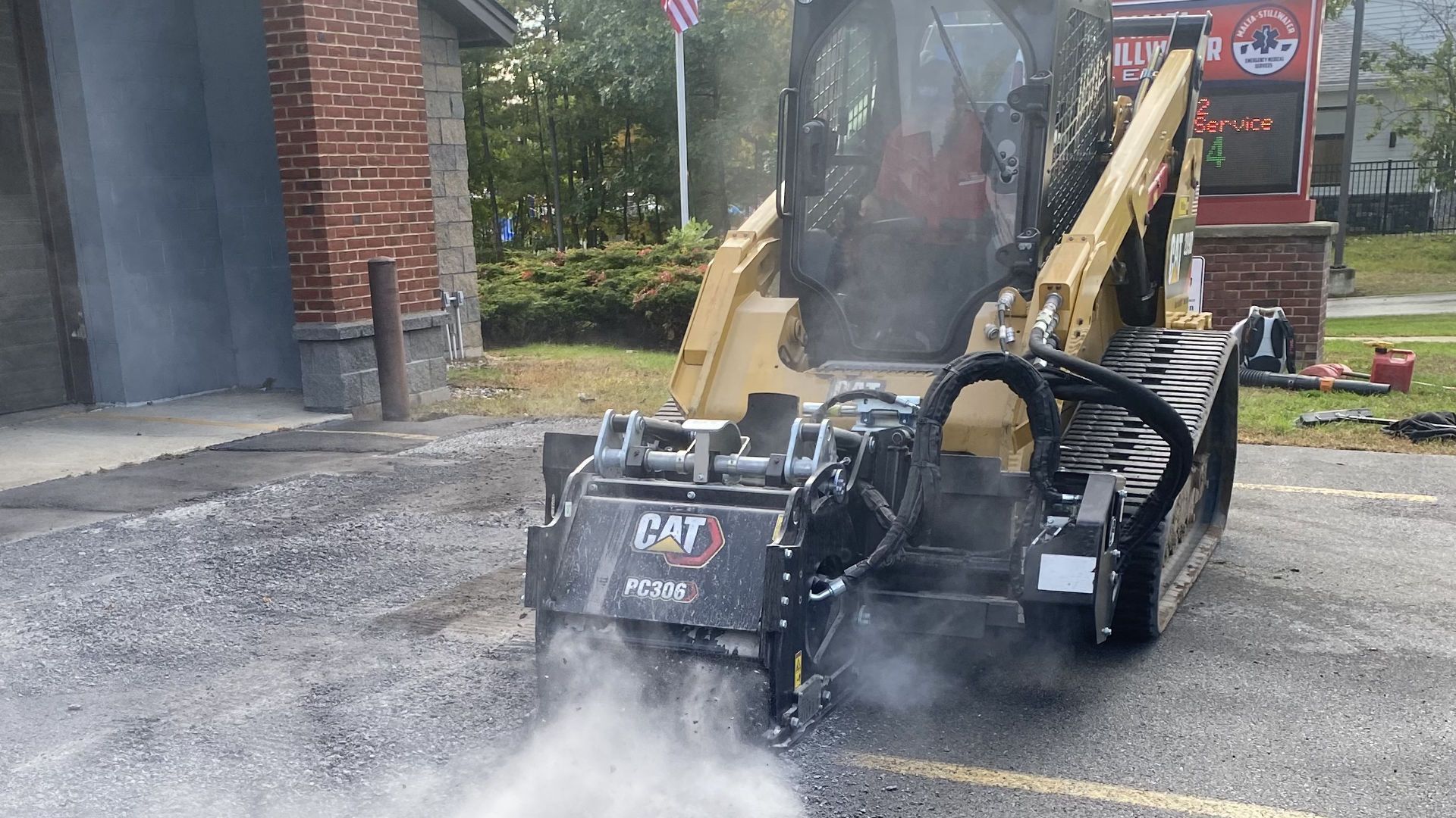 Yellow CAT skid steer with milling attachment grinding asphalt, creating dust outdoors.
