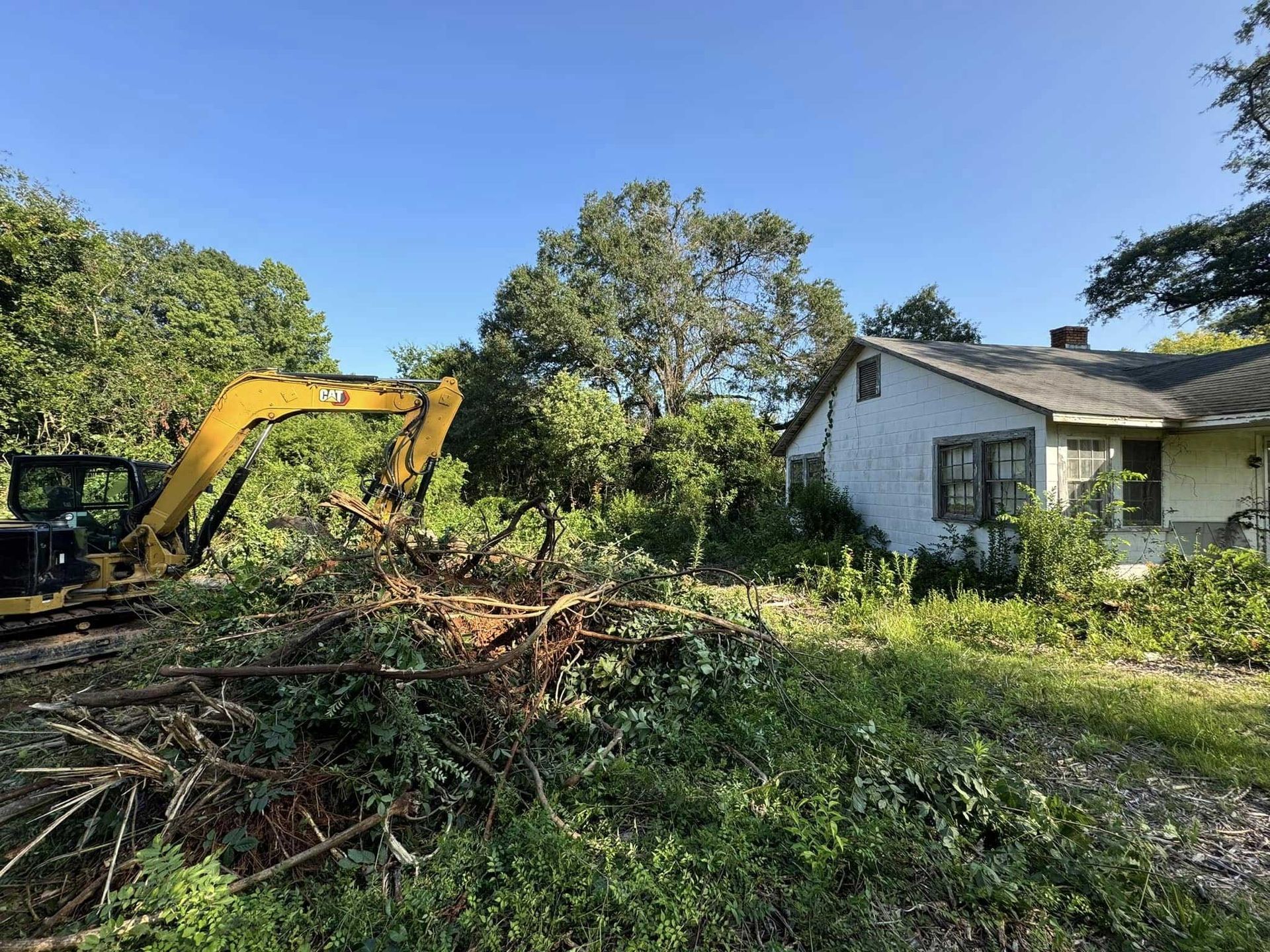 An excavator clears brush near a dilapidated white house with overgrown foliage under a blue sky.