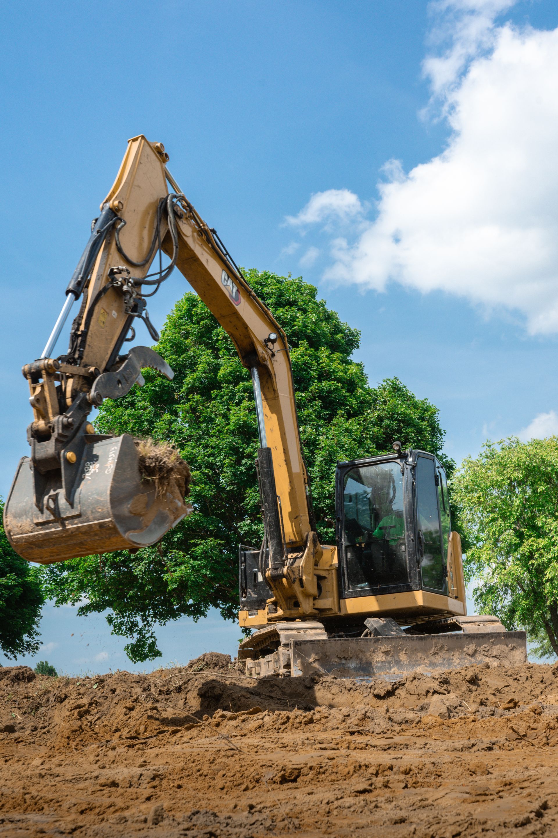 A yellow excavator works on a dirt construction site under a blue, cloudy sky with green trees in the background.