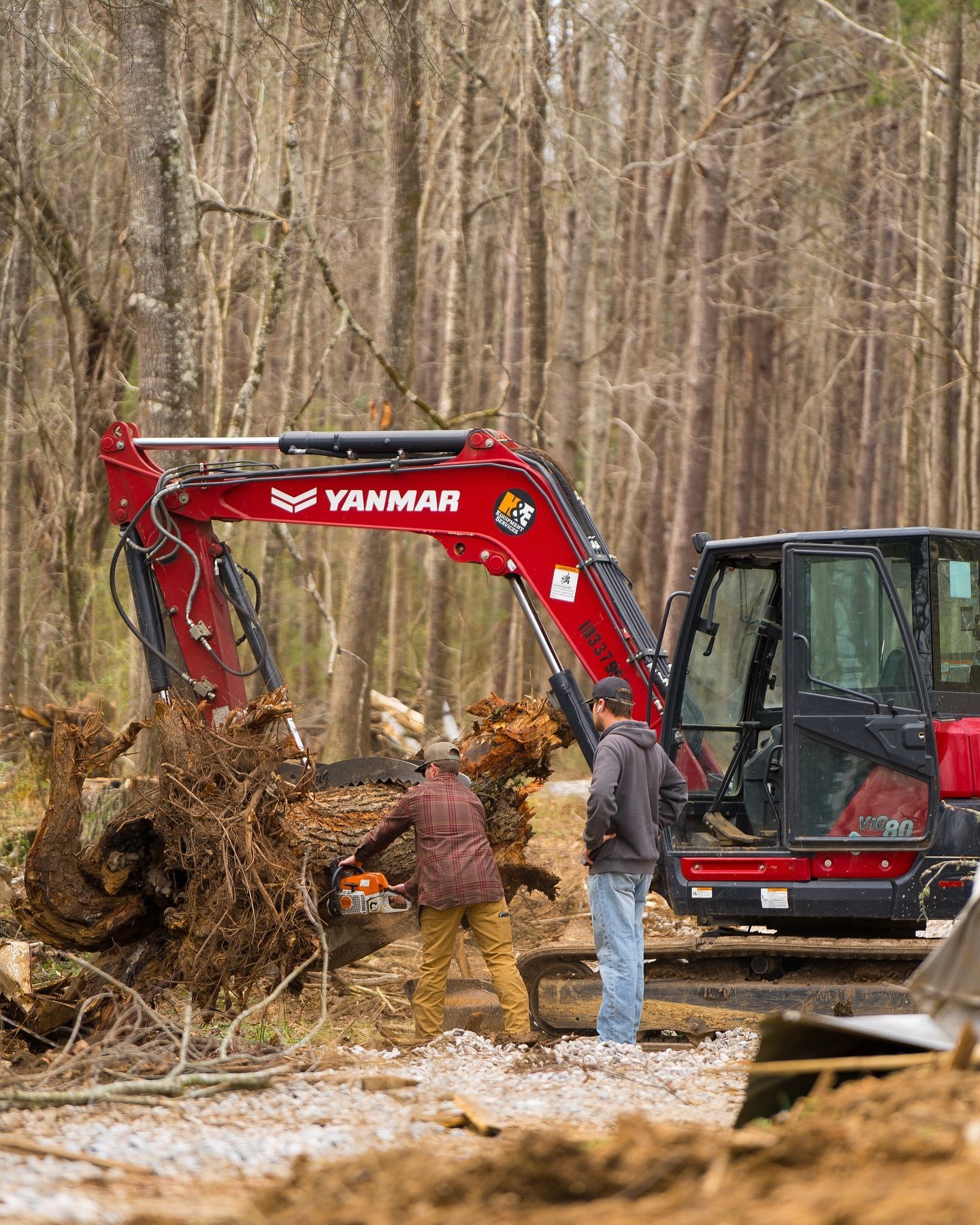 Red Yanmar excavator clearing roots; two men watch in a wooded area.
