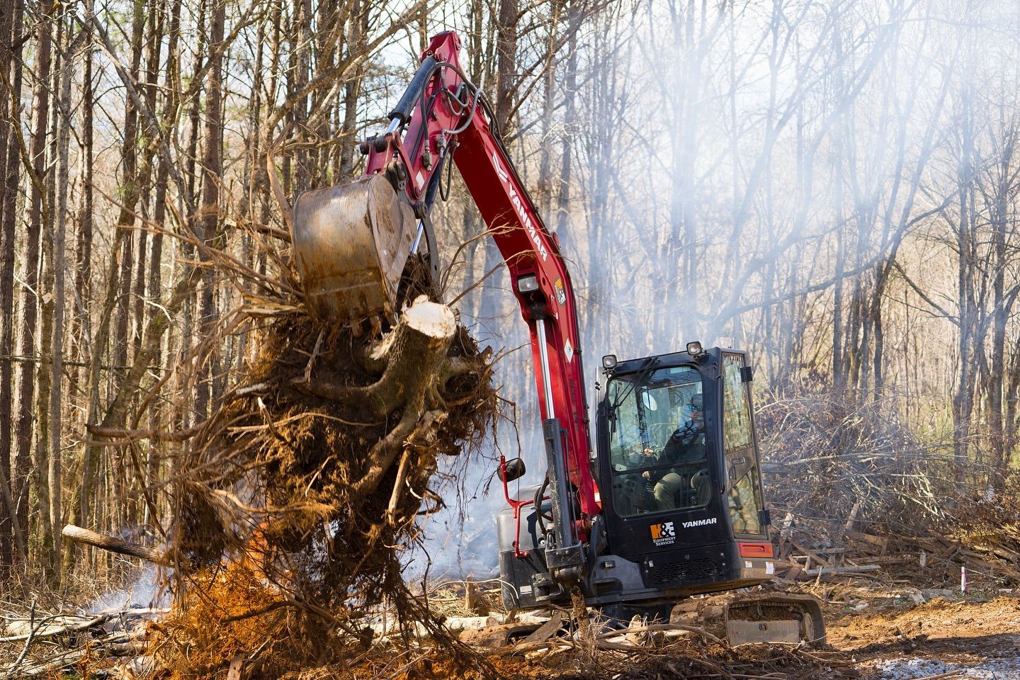 Red excavator uproots a tree stump in a forest clearing. Smoke rises in the background.