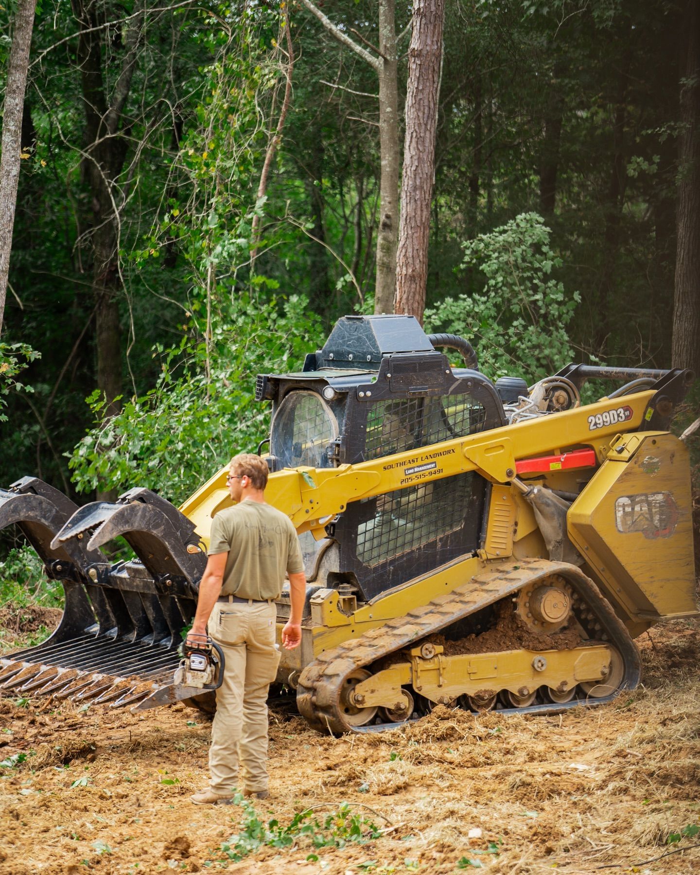 A person stands near a yellow Caterpillar skid steer with a forestry mulcher attachment in a wooded area.