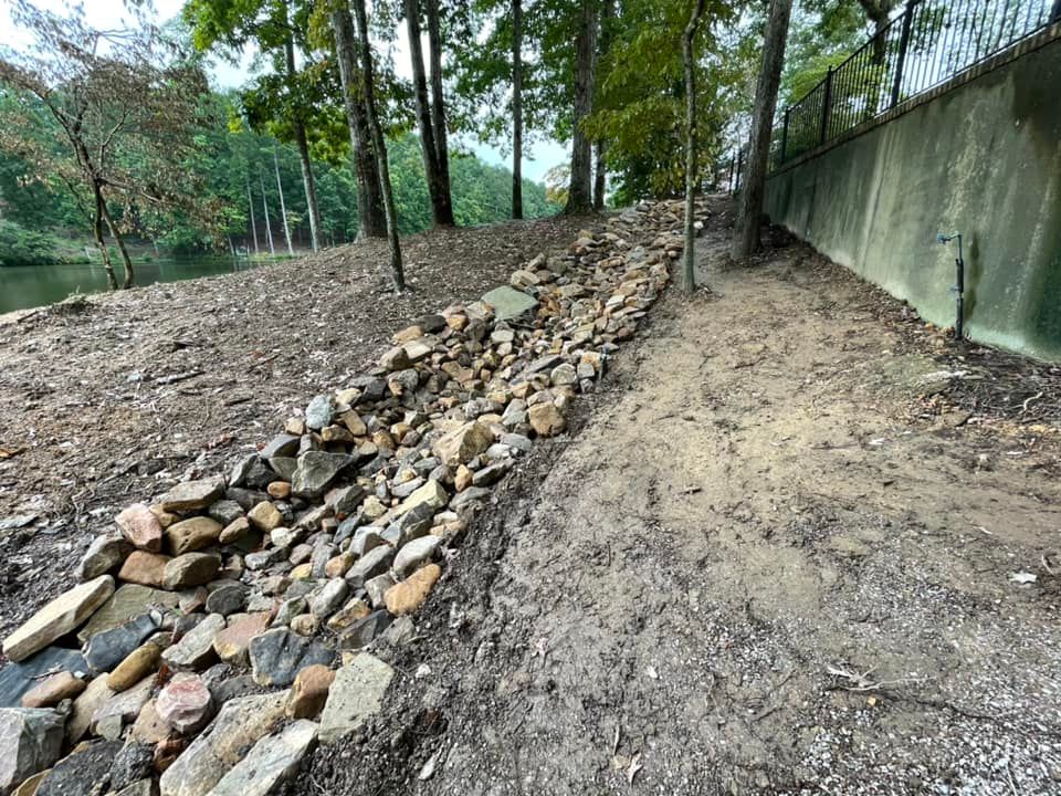 A stone drainage ditch runs alongside a concrete wall and a wooded bank near a body of water.