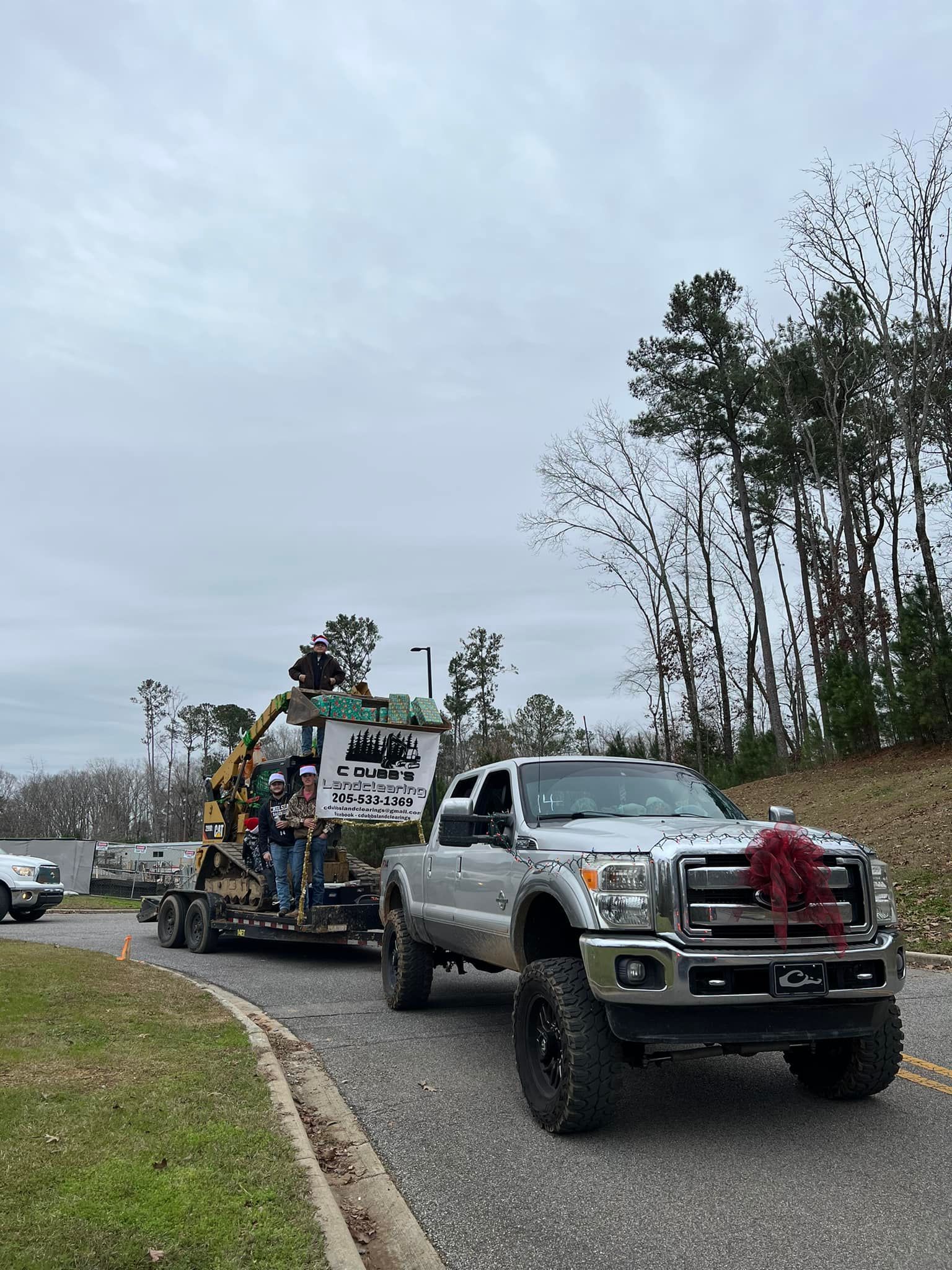 Silver pickup truck towing equipment on a trailer. A Christmas bow is on the truck's grill.