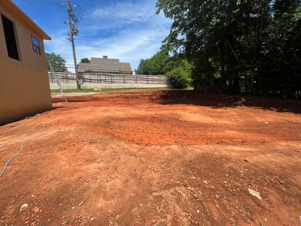 Cleared red dirt lot next to a tan building, with a few trees and a fence in the background.