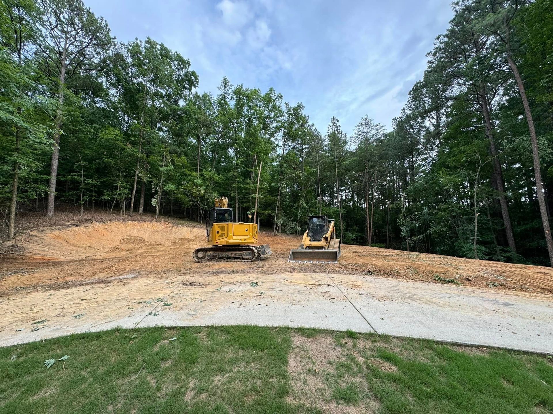 Construction site with yellow excavator and skid steer on a cleared plot, bordered by trees.