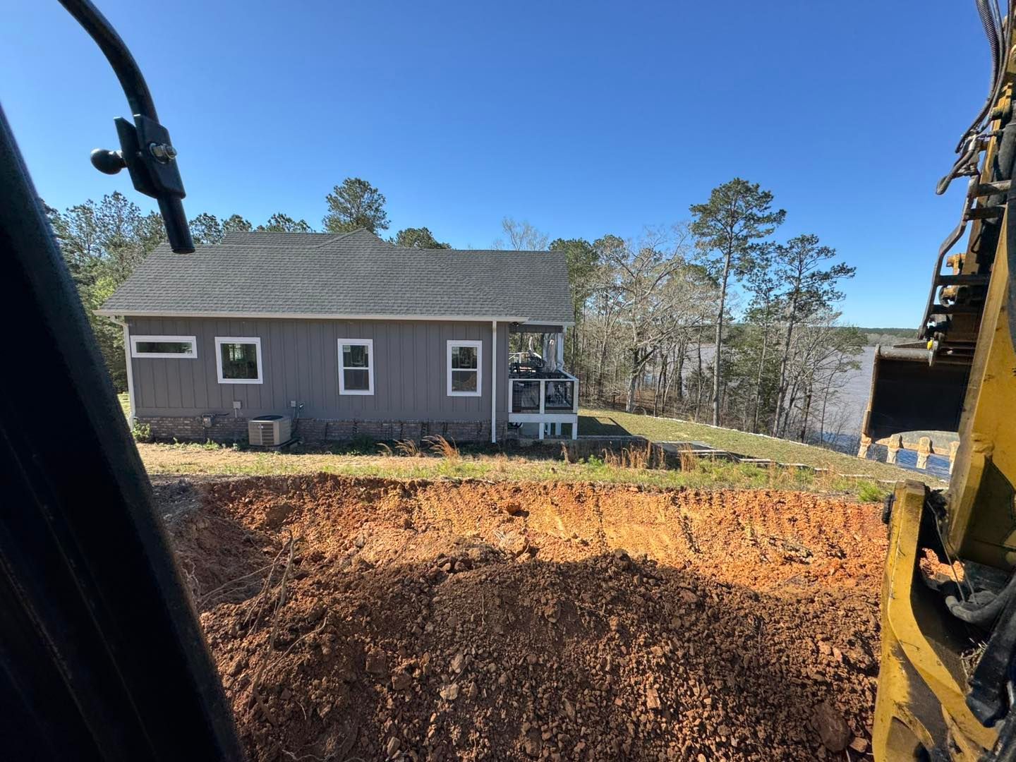 A house sits on a hill overlooking water, with an excavator in the foreground digging a trench.