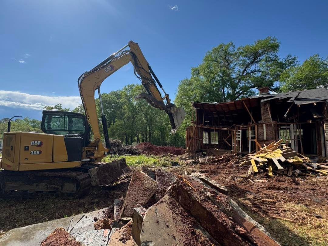 Yellow excavator demolishing a partially burned wooden house under a blue sky, with trees in the background.