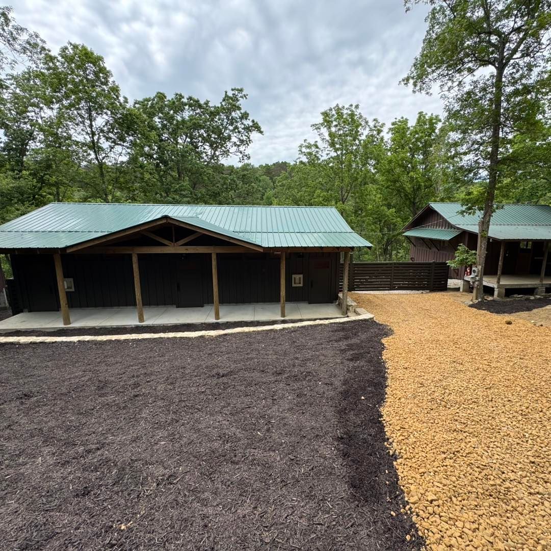Two rustic cabins with green roofs and wooden beams surrounded by trees and mulch.