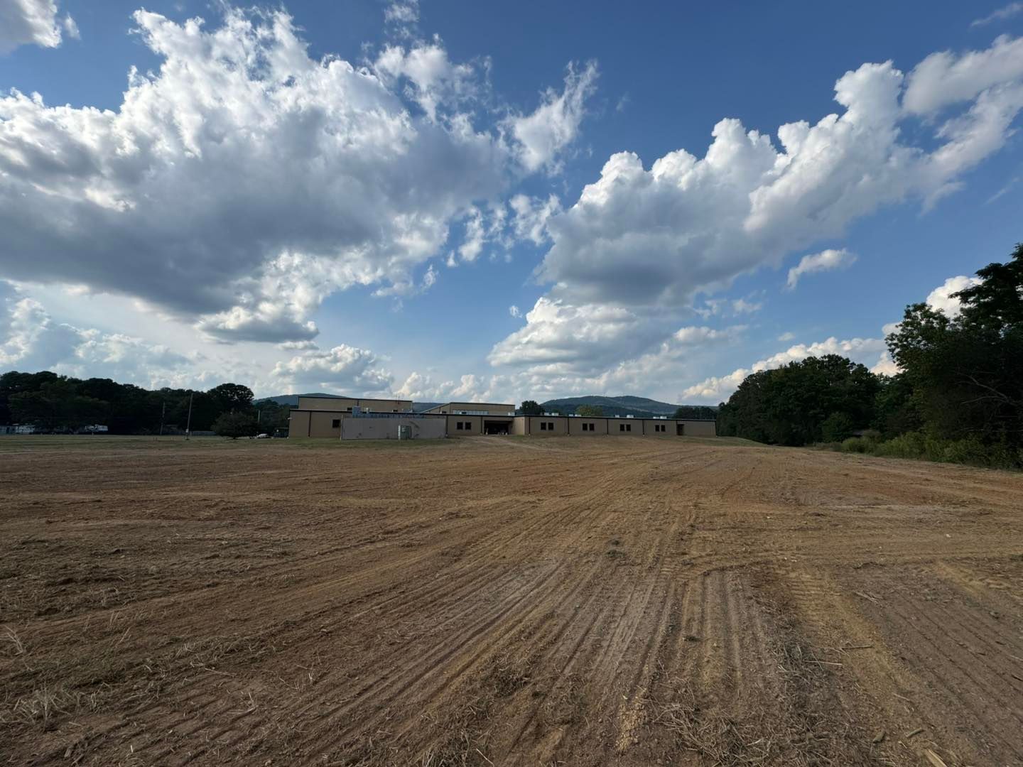 Cleared dirt field with a long, low building under a blue sky with clouds.