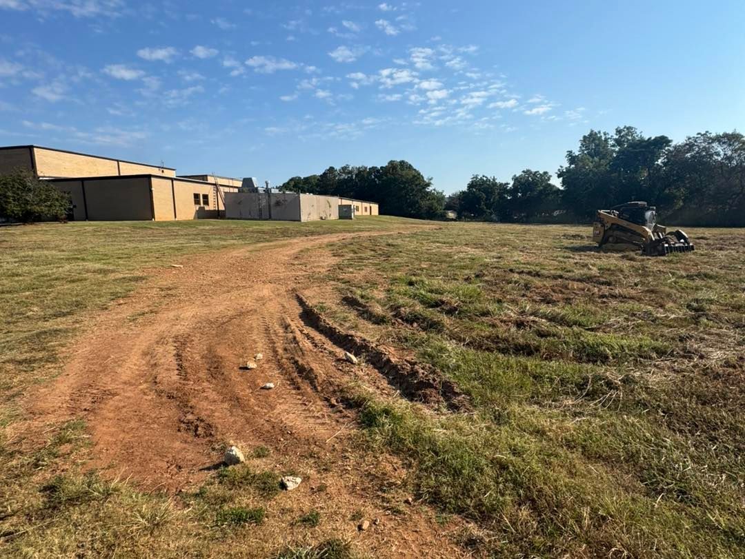 Dirt path through a grassy field, buildings in the background, blue sky.