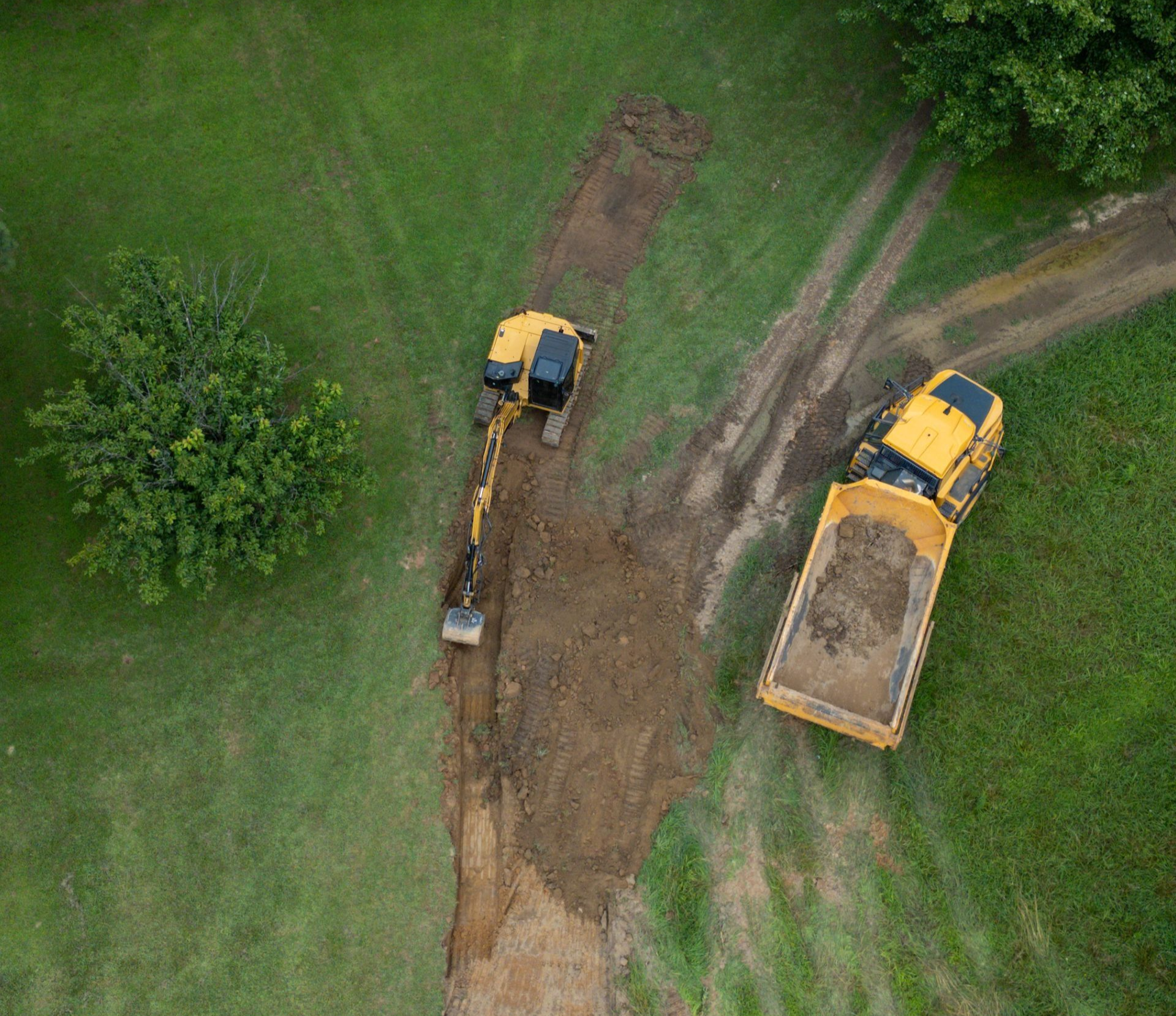 An aerial view of an excavator digging a trench in a grassy field next to a loaded yellow dump truck.