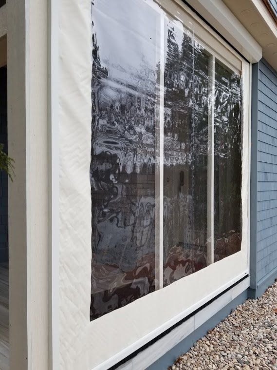 Clear vinyl outdoor roller blinds on a blue building, framing a patio or deck. Cream-colored trim.