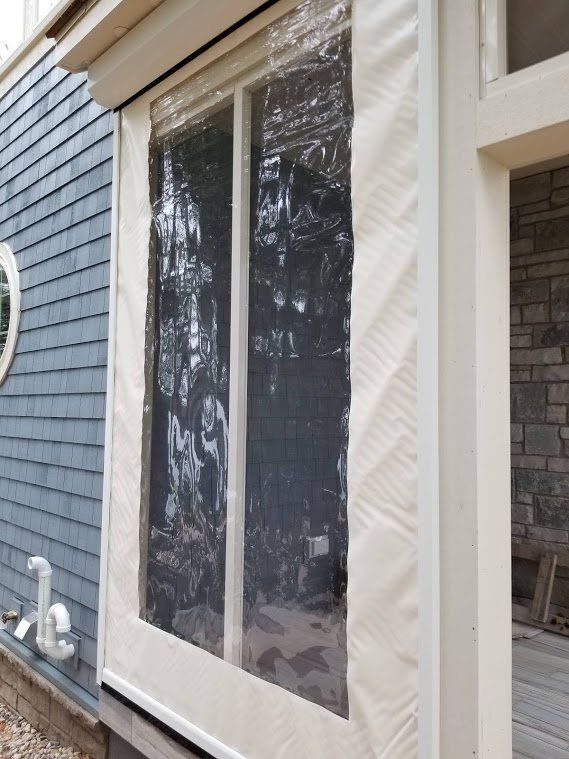 Clear vinyl and cream-colored fabric window cover on a house. The cover is rolled down over two window panels, with a stone wall to the right.