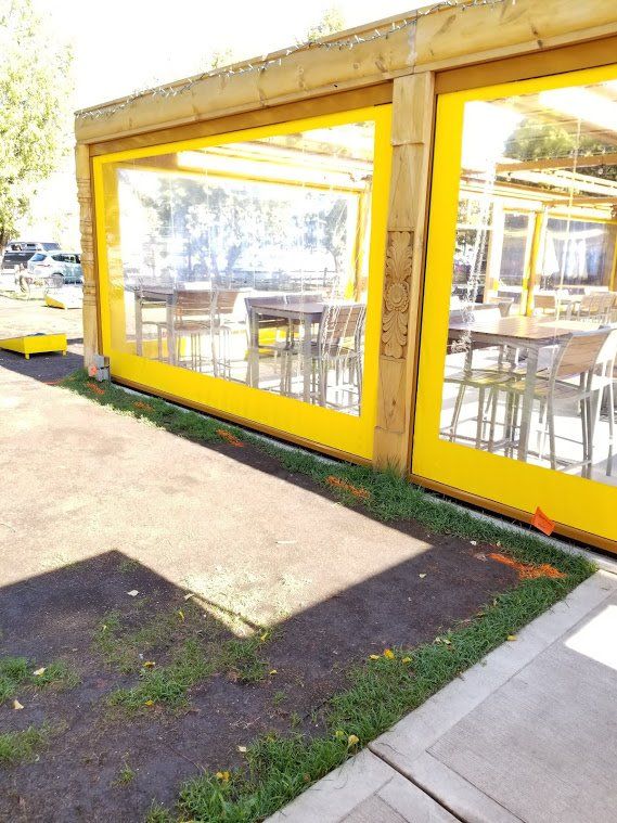 Outdoor dining area with yellow vinyl roll-down sides, tables, and chairs inside. The area is covered by a wooden pergola.