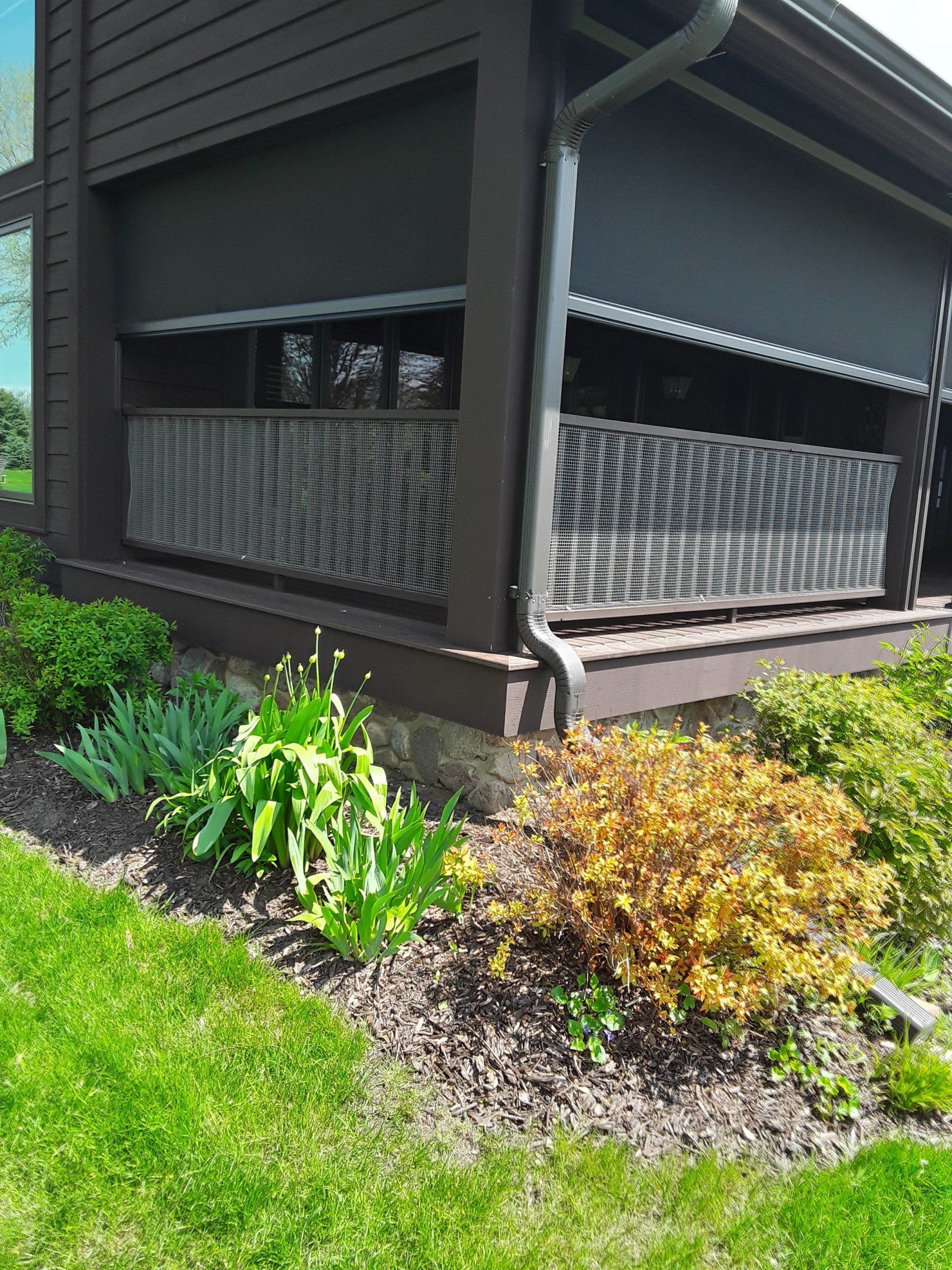Exterior of a house with a dark porch, sun shades, and a woven railing, surrounded by a garden with green plants and grass.
