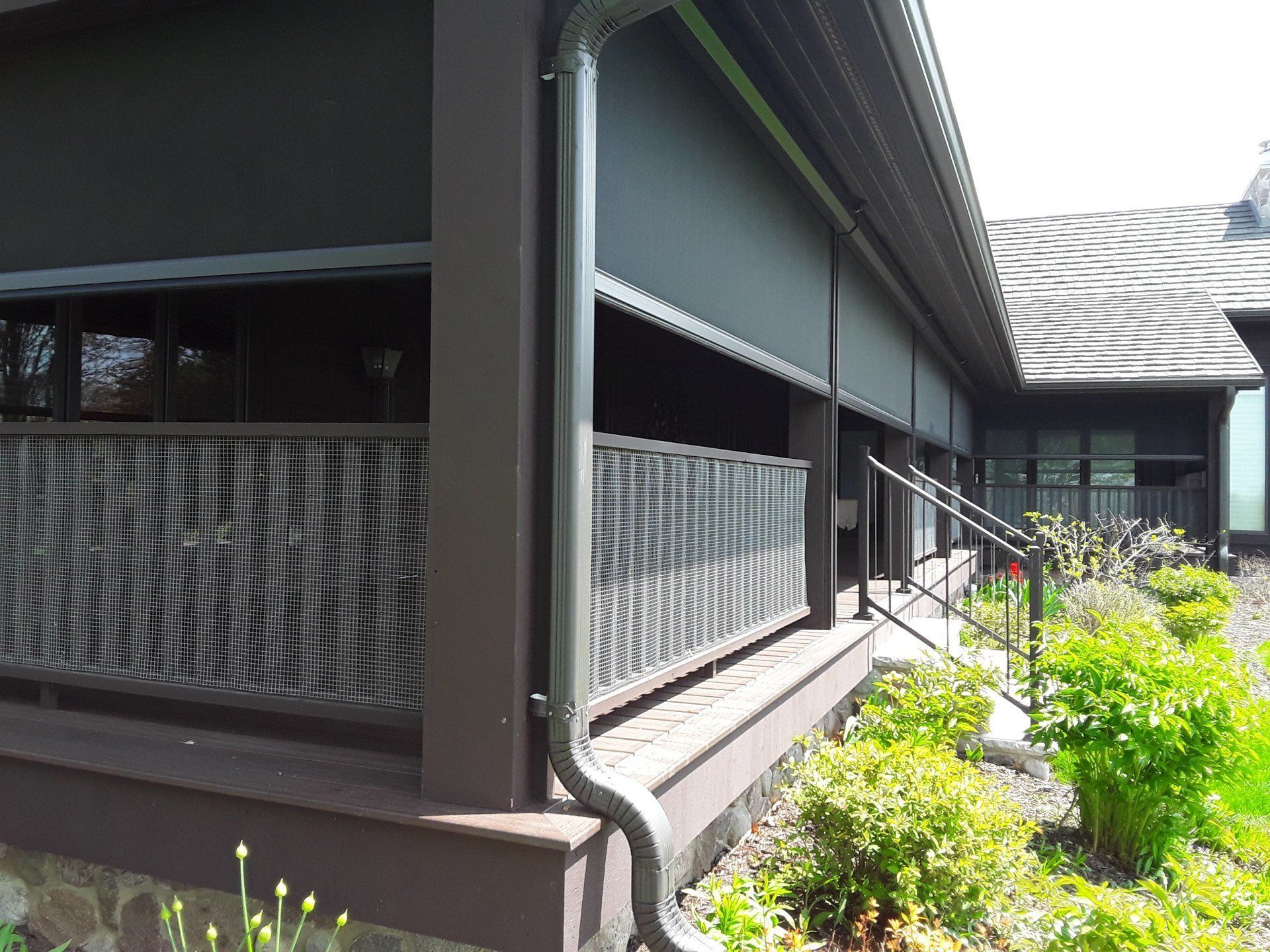 Brown outdoor retractable shades on a porch, with a metal railing and greenery.