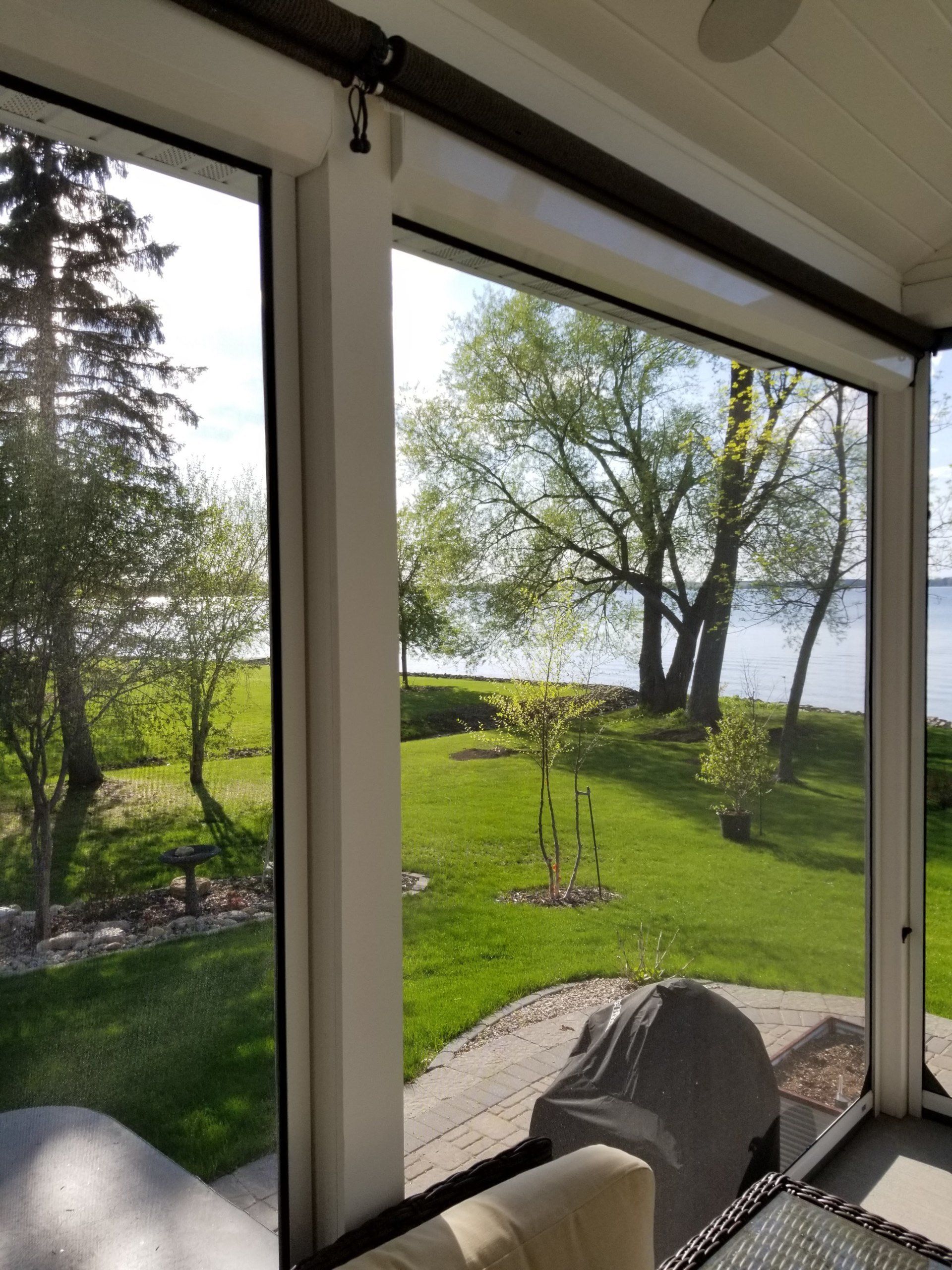 View of a green lawn, trees, and a lake through screened porch windows. The porch has white frames and dark brown blinds.