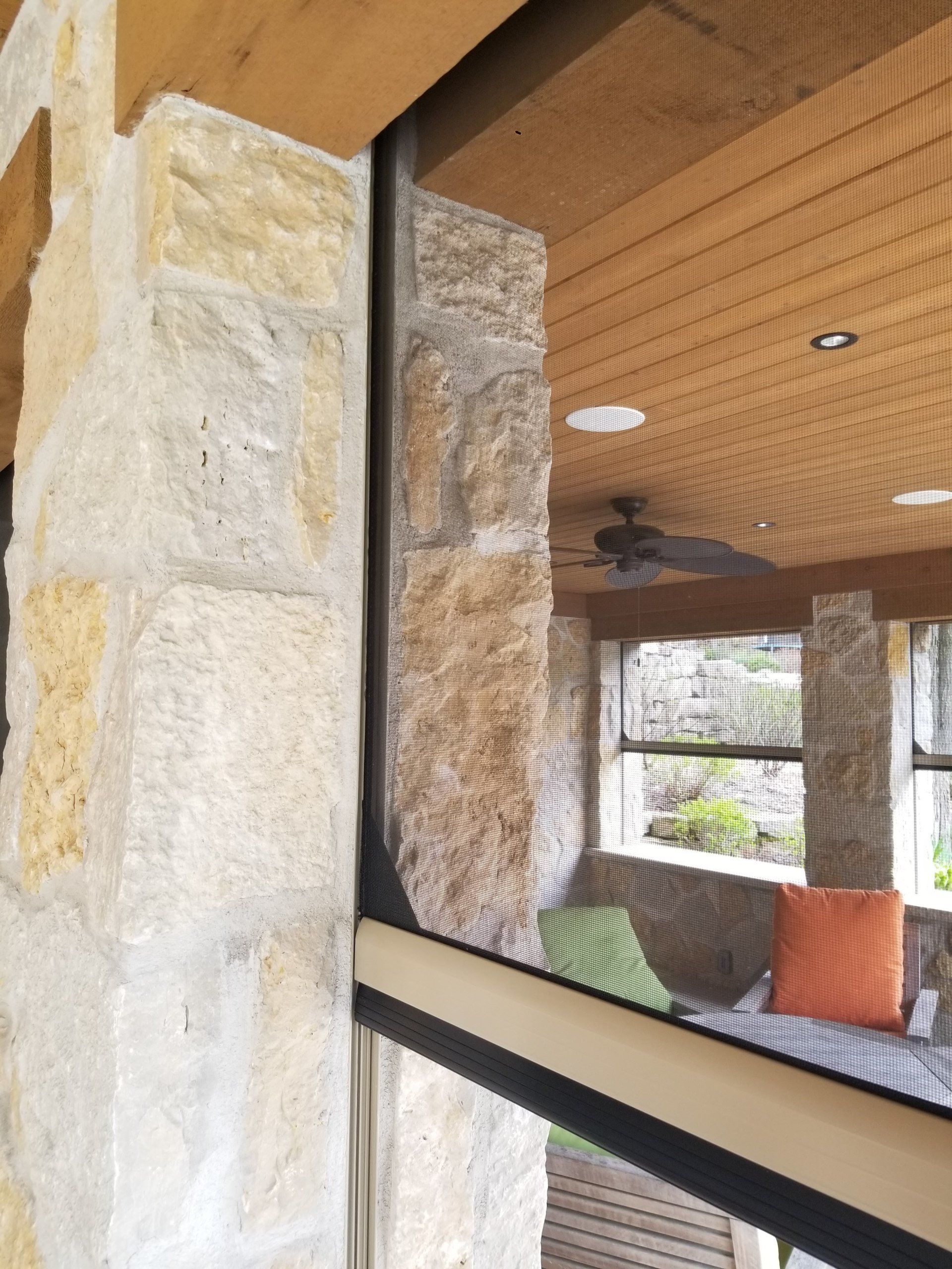 Close-up view of a window frame next to a stone pillar. The window reflects an outdoor living space with stone columns and a ceiling fan.