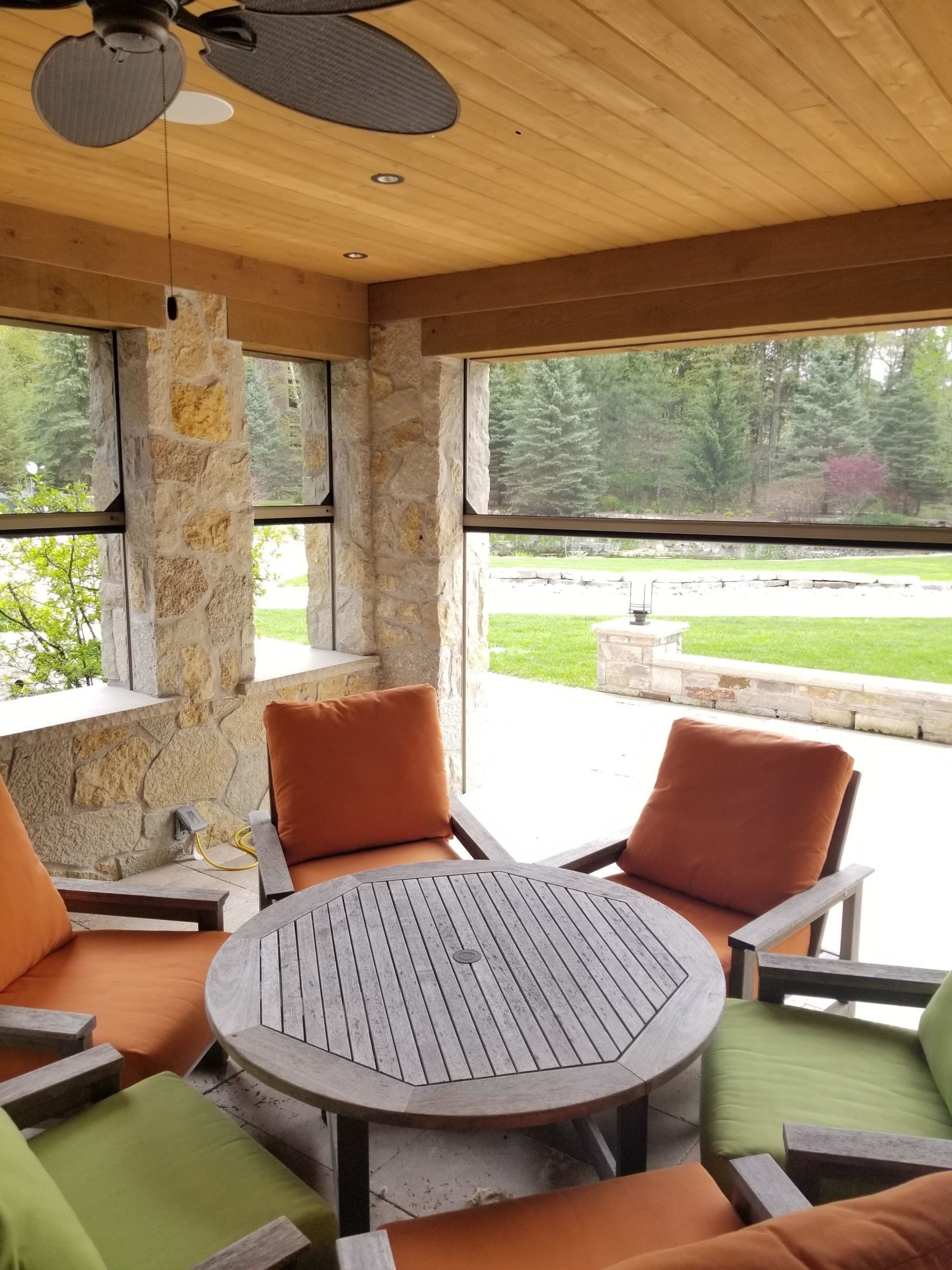 Outdoor seating area with stone columns, wood ceiling, ceiling fan, and chairs with orange and green cushions. Retractable screens are partially lowered.