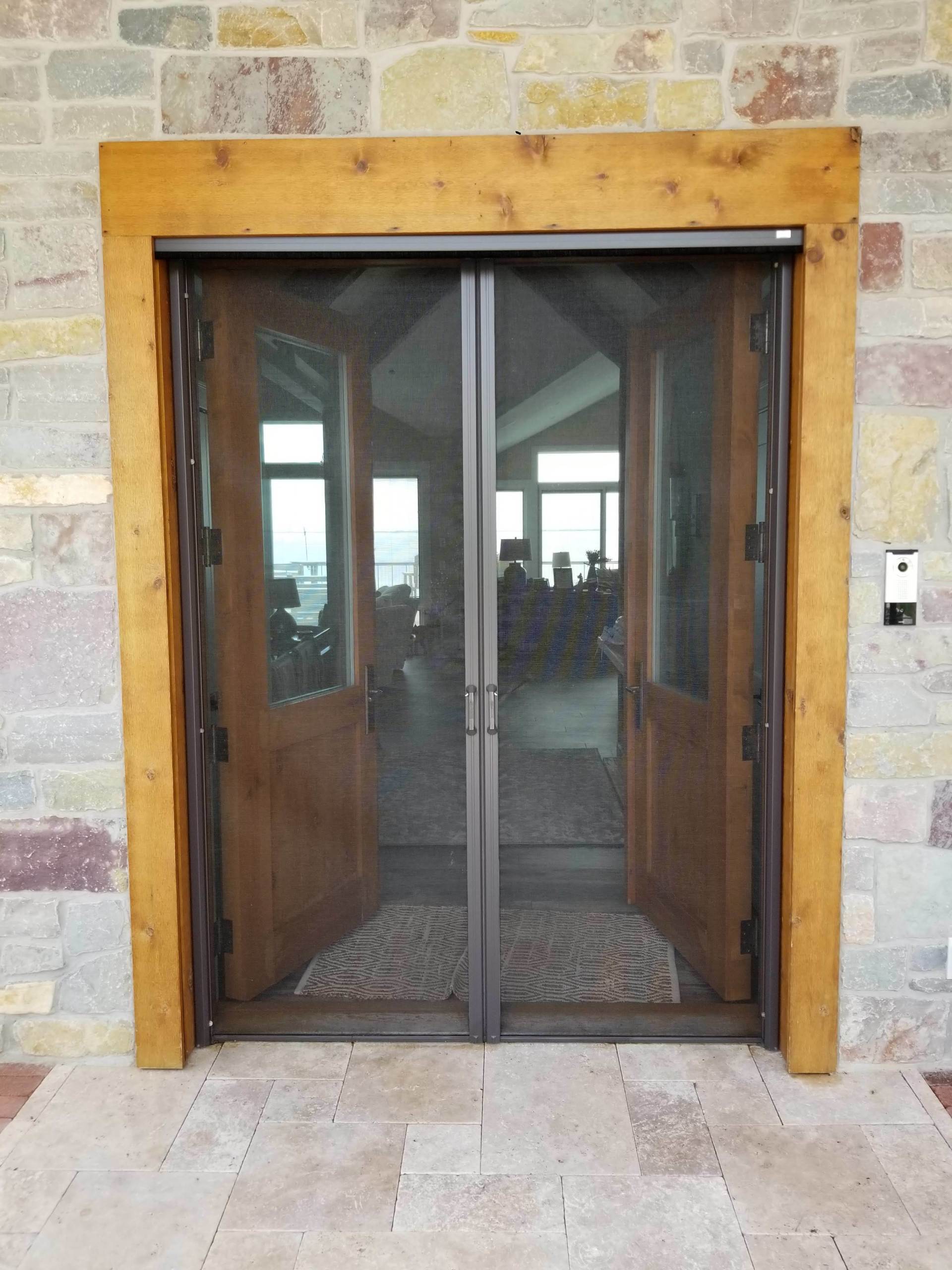 Double wooden doors with screen door, set in a wooden frame. The doors are open to a light-filled room.