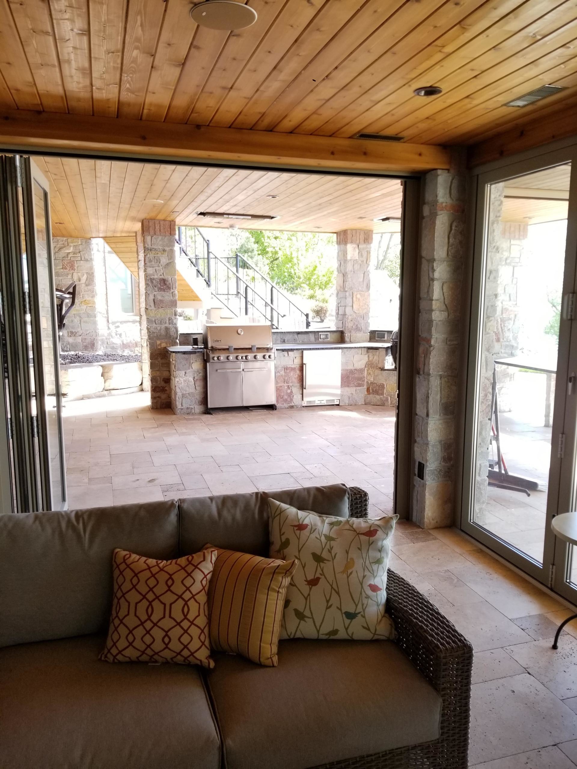 A cozy outdoor living space with a sectional sofa faces an open-air kitchen and grilling area; wood ceiling and stone columns frame the view.